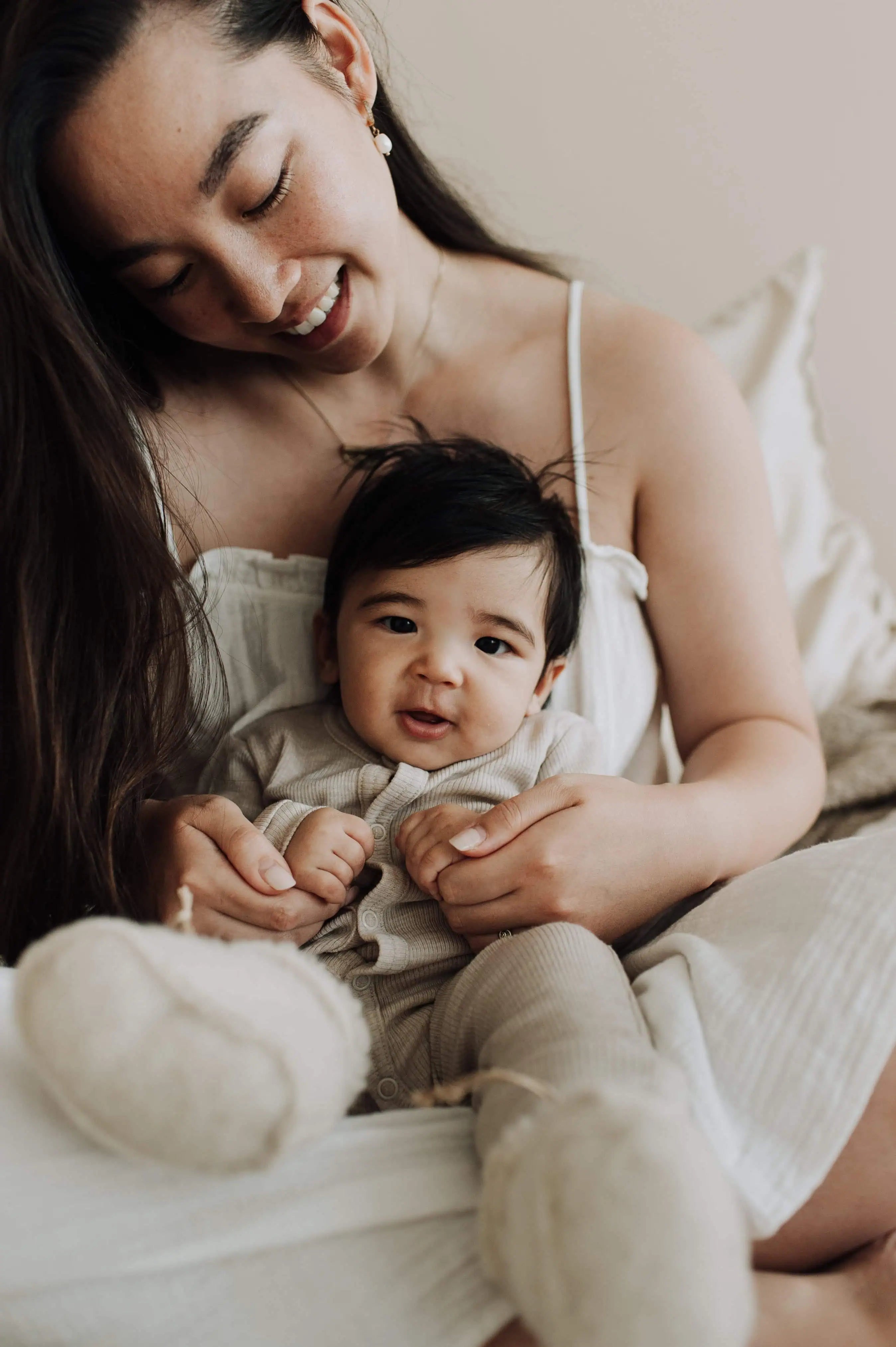 Moeder met zwart haar die met baby in wollen babykleding op schoot zit waarbij ze elkaars handen vasthouden