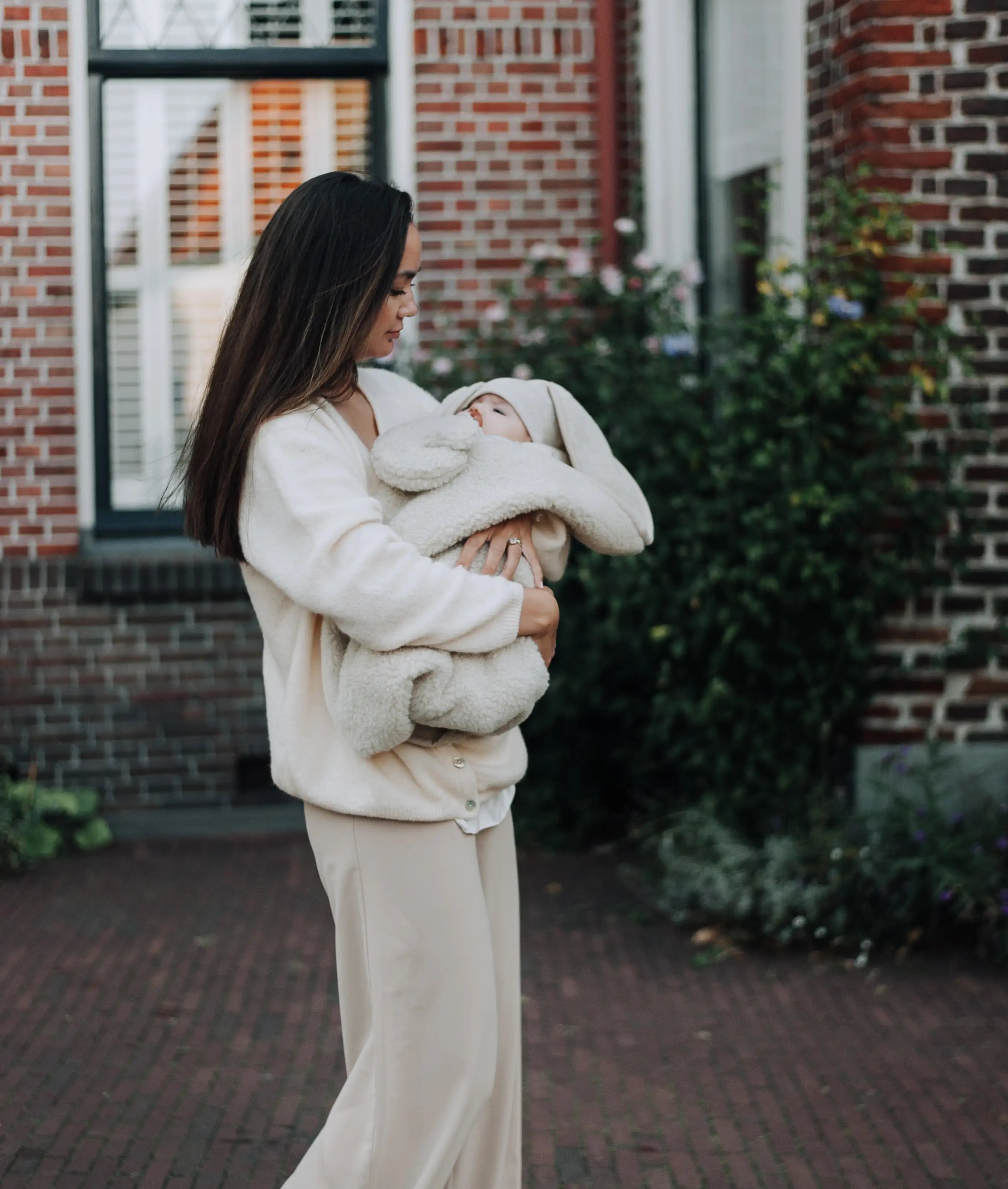 moeder die wandelt met baby in armen die gewikkeld is in een wollen deken. Het kind heeft een spin in met daaraan een wollen knuffel