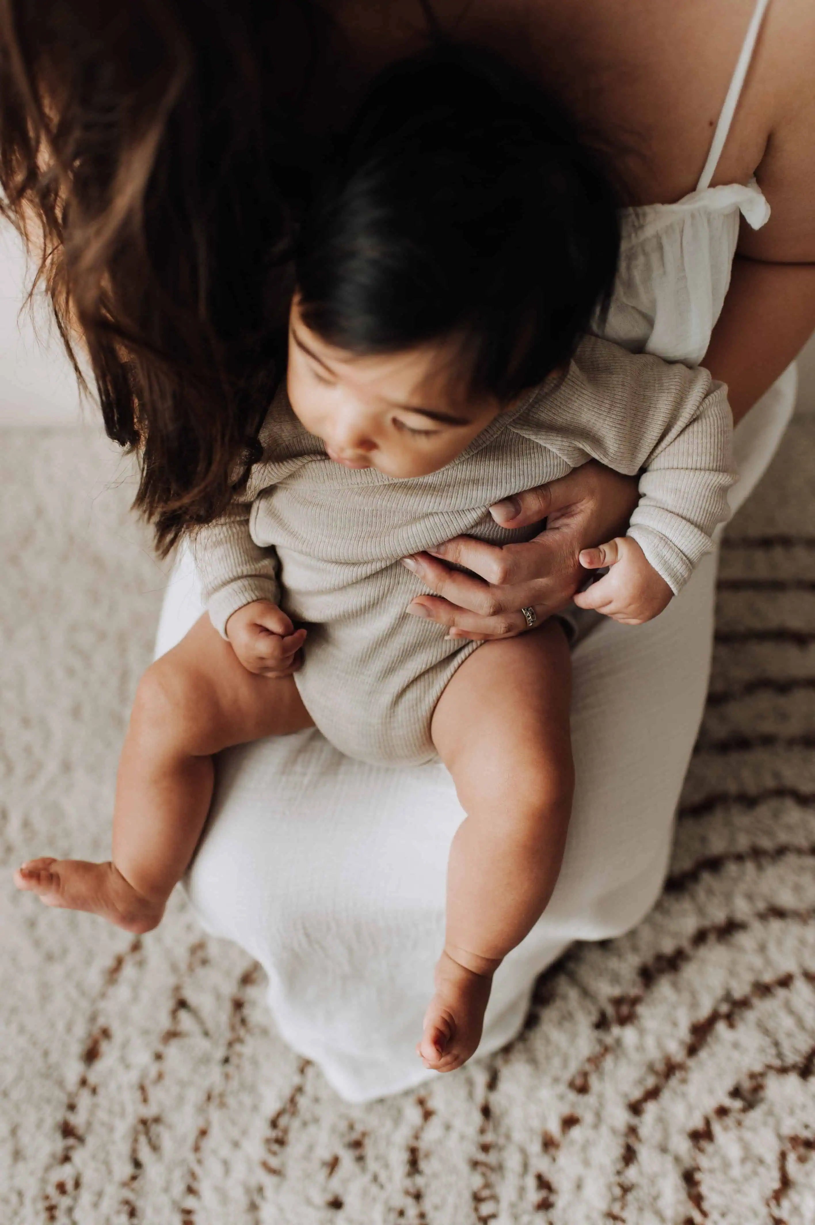 Baby met zwart haar in een merinowol overslag romper die bij zijn moeder op schoot zit. Van bovenaf geschoten